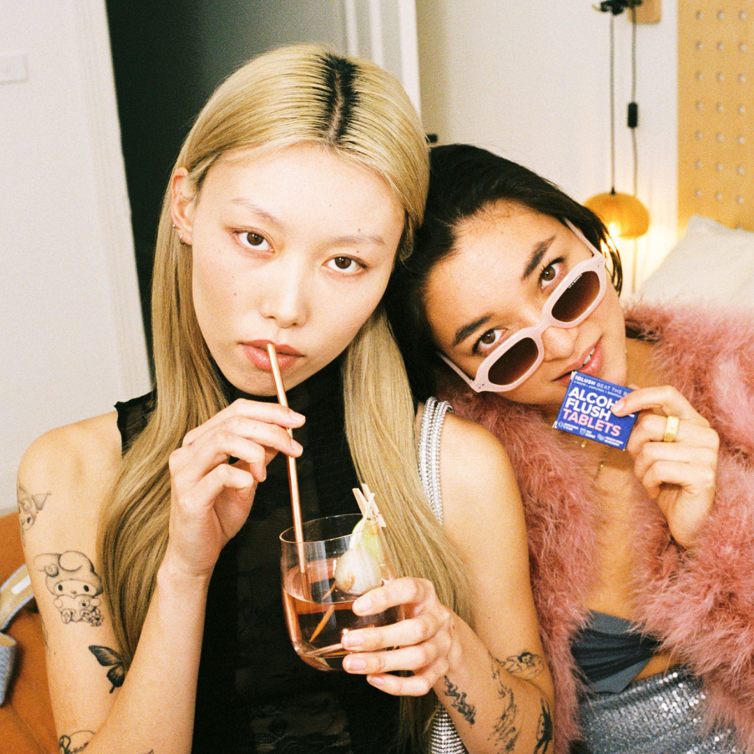 Two women posing together, one drinking from a glass with a straw and the other holding the iBlush alcohol  flush tablets packaging.