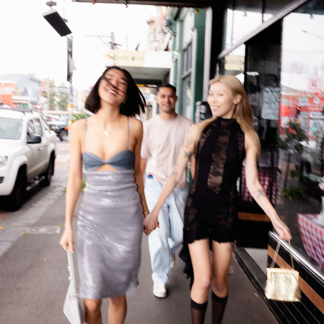 Two women walking on a city street, one in a silver dress and the other in a black outfit, with a man in the background.