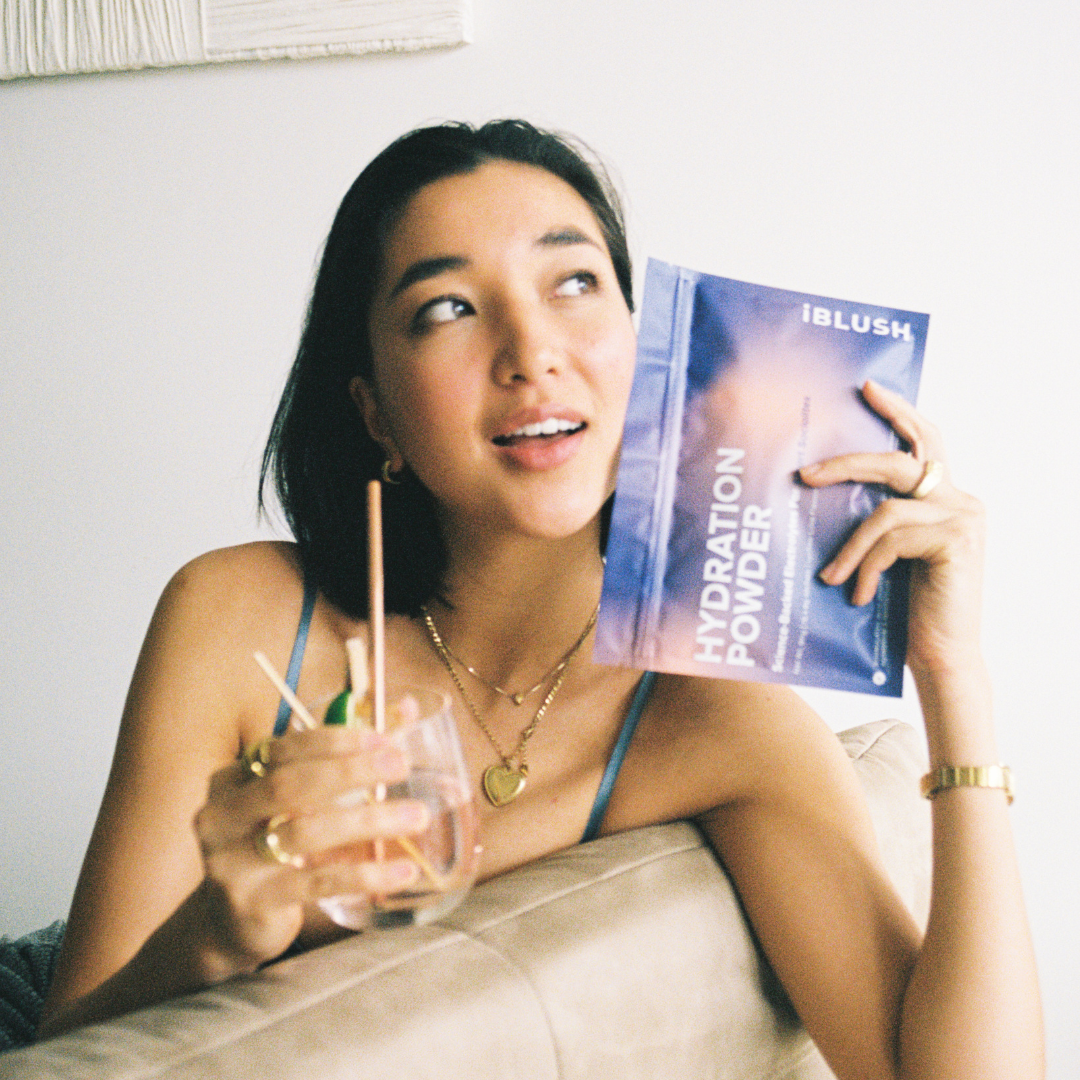 Woman on film holding iBlush Hydration Powder pouch, a sugar-free electrolyte powder designed to support hydration and recovery while drinking.