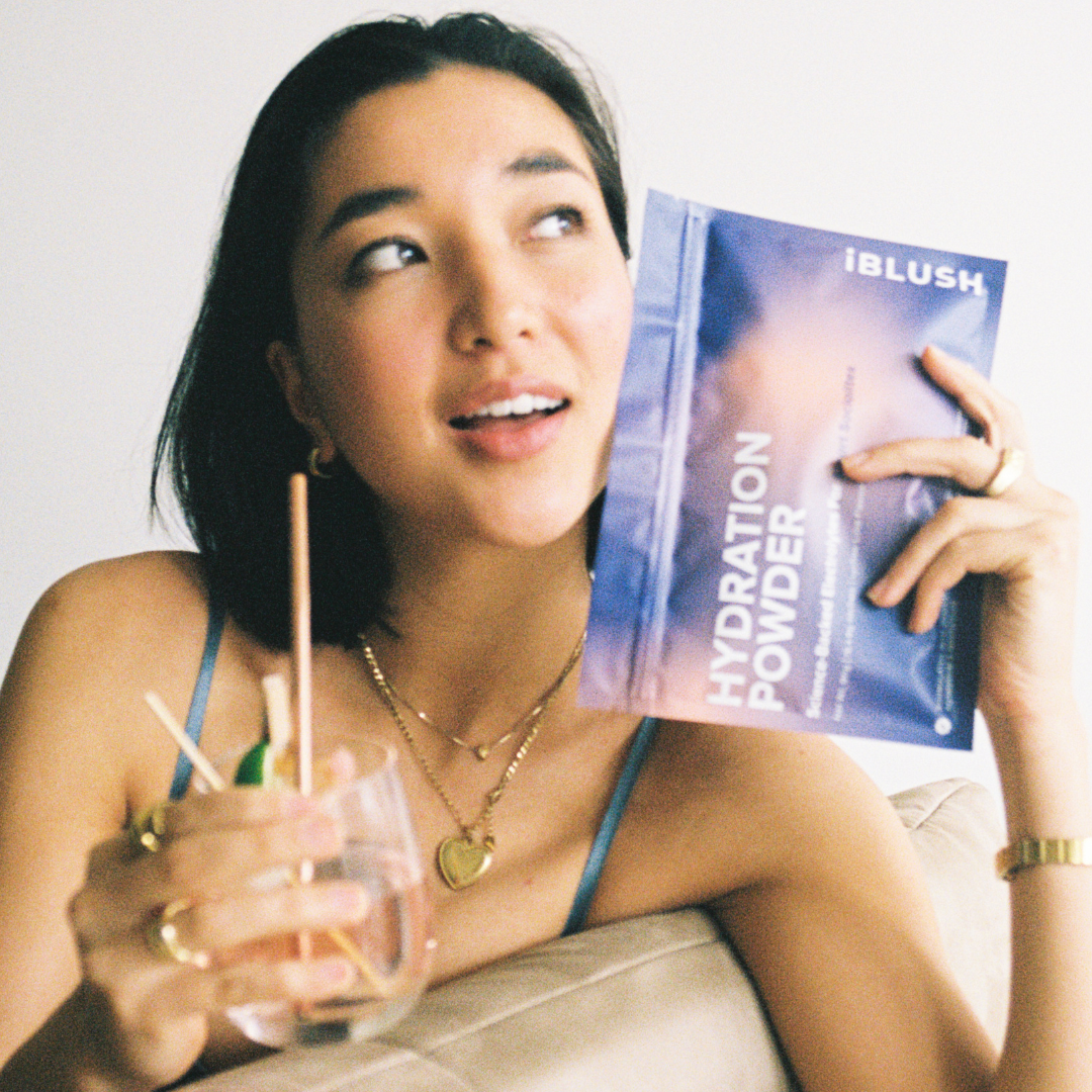 Woman on film holding iBlush Hydration Powder pouch, a sugar-free electrolyte powder designed to support hydration and recovery while drinking.